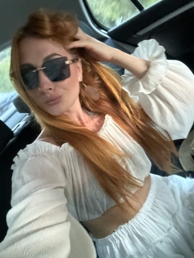 A woman is photographed in a selfie-style shot inside a vehicle in Prijevorac, Montenegro. She possesses long, reddish-blonde hair and is wearing large, dark, rectangular sunglasses with gold-colored embellishments. A subtle nose piercing is visible on her left nostril. She is dressed in a white two-piece ensemble featuring an off-the-shoulder, ruffled, pleated crop top with voluminous sleeves and a matching pleated skirt. Large, light brown, flat, ear-shaped earrings are present. Her right hand is positioned near the top of her head, touching her hair. The car's interior, including dark upholstery and a window revealing blurred green foliage, forms the background. The subject maintains a relaxed facial expression while looking towards the camera.