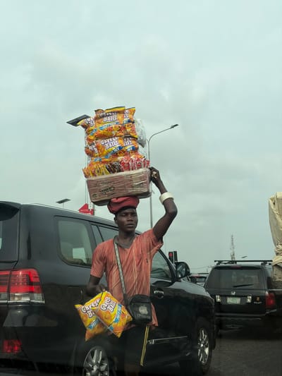 A male street vendor stands amidst stationary traffic on a road in Ikate, Nigeria, under an overcast sky. He is dark-complexioned, wearing a red and black striped shirt, black shorts, and a red head wrap. A white bangle adorns his left wrist, and a black "iPhone" branded crossbody bag hangs over his right shoulder. He carries a tall, rectangular stack of merchandise on his head, primarily consisting of large yellow bags labeled "Cheese Balls PARTY PACK," with smaller snack bags beneath, all secured by clear plastic. In his left hand, he holds two additional "Cheese Balls PARTY PACK" bags. His right arm is raised, with his hand near the top of the merchandise stack. To his left is a dark-colored SUV, likely a Toyota Land Cruiser, displaying its rear lights. Behind it and to the right, a black Honda Pilot is visible. A street light pole and a red traffic signal are present in the background.