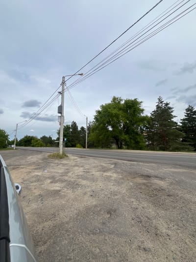 A roadside view in Malyshevo, Russia, shows a two-lane asphalt road with a gravel shoulder. Power lines crisscross overhead, supported by concrete utility poles. One pole, in the foreground, has a gray electrical box attached to it, along with two signs; one appears to be a bus stop sign. On the right side of the road, a dense line of trees, including tall evergreen trees and a large deciduous tree with green foliage, forms a backdrop. The sky is overcast with clouds. The left side of the image shows the edge of a silver car.