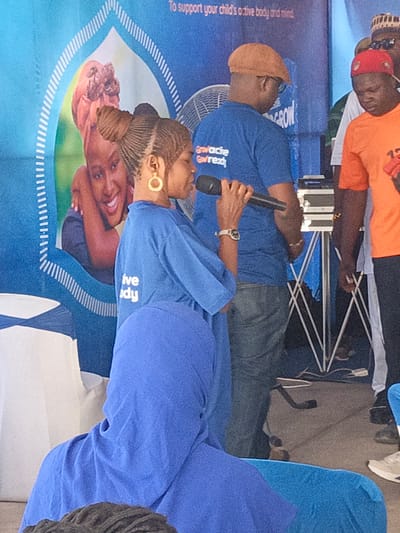 The image captures an indoor or covered outdoor event, likely a public speaking engagement or presentation in Tarauni, Nigeria. The central focus is a woman in a royal blue t-shirt, holding a black microphone to her mouth, suggesting she is speaking or singing. She has intricate braided hair styled into a bun and wears prominent gold hoop earrings. Her blue t-shirt has partially visible white text that reads "ive dy," likely part of a phrase like "active body."

Behind her, a man in a similar blue t-shirt and jeans is visible from the back. He wears a light brown flat cap, and his shirt has white text that appears to read "Gwache Gortez" or "Gwoche Gortex." To his right, another man in an orange t-shirt and a red cap is also seen from the back. A white chair back is visible to the left of the speaker, and a person in a blue hooded garment is partially visible in the foreground, sitting on a blue surface.

The background features a large blue banner with white arch-like designs. An image of a smiling woman embracing or holding a child is prominent on this banner. Visible text on the banner's top right corner states: "To support your child's active body and mind." A white fan is positioned behind the man in the blue shirt, and some audio equipment on a stand is visible to his right. The overall atmosphere suggests a promotional or community event focused on health or development, given the banner's message and the coordinated attire.