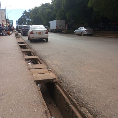 The image shows a street scene in Sheywe ward, Kenya. A drainage ditch runs along the left side of the asphalt road, covered intermittently with concrete slabs. The ditch contains debris and stagnant water. Several cars are parked or moving along the road, including a white sedan with license plate KBU 104H. A truck is also visible further down the street. People are present on the sidewalk on the left side of the road, near a Coca-Cola sign. Trees line the background.