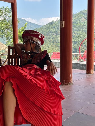 The image, rotated 90 degrees clockwise, portrays a person in traditional attire resting on an outdoor terrace or balcony, overlooking a mountainous landscape in Thông Nguyên Commune, Vietnam.

The central subject, likely a woman, is shown from the waist up, leaning forward on a wooden chair with her head gently bowed. She wears a striking traditional costume featuring a rich red and silver headpiece, intricately adorned with metallic plates and beads. Her upper garment is a black, long-sleeved top with vibrant, colorful embroidery. The most prominent part of her attire is a voluminous, pleated red skirt that flows downwards, creating a dramatic visual impact. One hand, with long, light-colored manicured nails, is visible, resting on the skirt.

The setting appears to be a raised outdoor area, possibly a viewing platform or balcony. The floor is covered with reddish-brown tiles, laid in a diamond pattern. Sturdy wooden columns and railings frame the space; an electrical outlet is visible on one of these columns. Beyond the terrace, a breathtaking view of lush green mountains under a bright, sunny sky unfolds. A partial glimpse of a red metal structure, possibly a chair or part of an architectural feature, can be seen in the background. The clear sky with some white clouds suggests a pleasant daytime scene. The image captures a serene moment, highlighting cultural heritage against a beautiful natural backdrop. No visible text is present.
