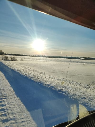 The image depicts a snow-covered winter landscape, viewed from inside a vehicle in the city of Малая Уса, Russia. The perspective is through a windshield or side window, with a dark brown vehicle frame visible at the top and a dashboard reflection in the lower right. A bright, unclouded sun is prominent in the upper left, casting strong rays across a clear blue sky with faint white clouds. The foreground and midground consist of expansive, flat snow-covered fields. A deep blue shadow extends diagonally from the lower left towards the center. Subtle tire tracks or disturbed snow are visible in the immediate left foreground. Sparse, leafless bushes dot the snow on the left side. A distant, dark treeline of what appears to be evergreen forest spans the horizon. Beyond this treeline on the far right, another snow-covered expanse is visible, possibly a frozen body of water or further fields, with a thin vertical dark object, possibly a utility pole, rising above the distant horizon. The vehicle's window exhibits multiple specks of dirt, smudges, and a thin vertical piece of debris in the mid-right portion, along with a prominent yellowish circular smudge in the lower right. No individuals or active subjects are present. The scene is brightly illuminated by direct sunlight.