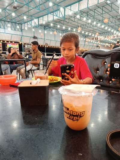 This image depicts an indoor dining area, likely a food court or restaurant, with a visible "Bintang Kopi" logo on a beverage cup in the foreground. The environment features a textured, dark tabletop, a black tufted leather chair, and a wooden tissue box. In the background, a young girl is seated, holding a smartphone with a decorative case. To her left, another person wearing a hijab is also looking at a phone. Further back, a man in a brown shirt is seated at a table. The ceiling structure is visible, with numerous lights and decorative hanging fixtures. The setting appears to be a casual, possibly outdoor-covered dining establishment. The presence of a "Bintang Kopi" branded cup suggests it could be a common or chain establishment. While the specific location within Namplohbaro, Indonesia is not explicitly stated, the visual cues point to a public eating area.