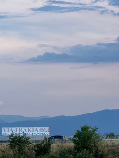 The image shows a tall, vertical billboard with text that appears to be a business advertisement. The billboard is positioned on the right side of the frame, set against a backdrop of rolling hills and a pale blue sky with wispy clouds. The scene suggests an outdoor, possibly rural or semi-rural location. A portion of a road and some green trees are visible near the billboard. The lighting and sky indicate it's daytime, likely late afternoon or early evening given the soft light. The text on the billboard is primarily in English and Cyrillic. The English text reads "VIA TRAKIA" in large letters and below it, "CASH & CARRY FOOD & NON FOOD". Above "VIA TRAKIA" in Cyrillic, the words "СКЛАД НА ЕДРО" are visible. The billboard is the most prominent object, serving as the focal point of the image. The surrounding natural landscape provides a serene contrast to the commercial advertisement. There are no people visible in the frame.