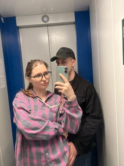 The image depicts a young couple taking a mirror selfie inside an elevator. The woman, in the foreground, is wearing glasses and a pink and grey plaid shirt. She is holding a smartphone, angled to capture the reflection. To her right, a man wearing a black baseball cap and a black jacket stands behind her. His left arm is around her waist. The elevator walls are a light grey with blue accents. The scene appears to be in an urban environment, possibly in Kirovo-Chepetsk, Russia, given the context provided.