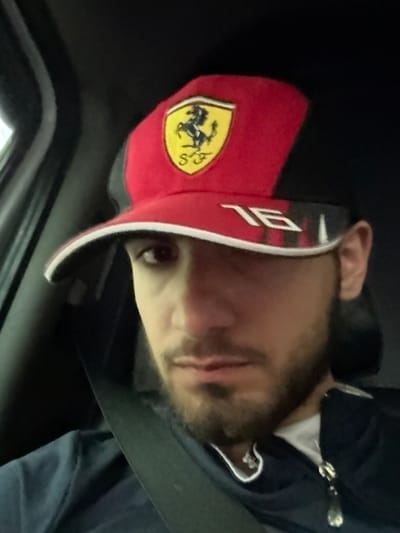 The image depicts an adult male seated inside a vehicle. He wears a red and black cap featuring the yellow Ferrari Prancing Horse logo with "SF" text, and the number "16" on the brim. His face is partially obscured by the cap, revealing dark eyes and a beard. He is wearing a dark blue zippered hoodie over what appears to be a white undershirt, with a black seatbelt fastened across his chest and left shoulder. The vehicle's interior is dark, with a portion of the door frame visible on the left side of the frame. The individual is looking directly towards the camera. The precise location within Tetri Tsklebi, Georgia, is not visually identifiable from the image.