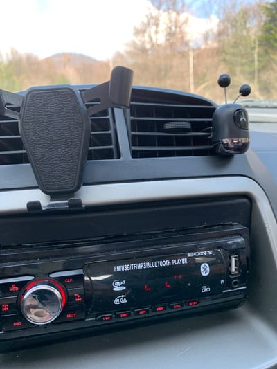 The image captures a detailed view of the interior of a car, specifically the dashboard area from a slightly tilted perspective. The main subjects are various car accessories and features.
In the center of the frame is a black Sony car stereo unit, labeled "SONY FM/USB/TF/MP3/BLUETOOTH PLAYER." It features a USB port, an AUX input, and various buttons, some of which are illuminated in red. Notable buttons include "CLK" (Clock), "MUT" (Mute), "MOD" (Mode), "BND" (Band), along with numbered preset buttons "1/||", "2/INT", "3/RPT", "4/RDM", "5/10-", and "6/10+". Symbols for Bluetooth, USB, IR, and MP3 are also visible. The stereo appears somewhat dusty.
To the left of the stereo, integrated into the air vent, is a black phone holder with a textured surface, designed to grip a smartphone. Above this holder, also attached to an air vent slat, is a small, black decorative item resembling an insect with two spherical antennae, possibly an air freshener or diffuser.
The background, visible through the car's windshield, shows a blurred outdoor scene with trees and bright sky, indicating it is daytime. The overall setting is inside a vehicle, likely in Gintsi, Bulgaria, as per the location context. No specific activity is occurring; the image is a static shot of the car's interior.