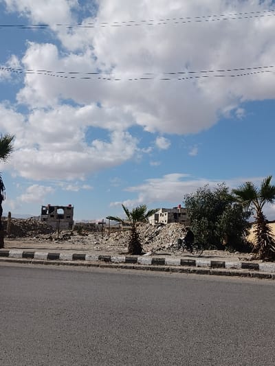 This image depicts a damaged urban environment in Al-Qadam Municipality, Syria. The foreground shows an asphalt road separated by a grey and black painted curb from a wide expanse of rubble and demolition debris. This debris forms uneven mounds across the midground. Several small palm trees are interspersed within the debris, alongside a large green bush on the right side. In the background, multiple partially constructed or damaged concrete buildings with exposed frames are visible, suggesting either unfinished development or structural ruin. One two-story concrete frame structure is prominent towards the left-center. An individual on a bicycle, dressed in dark clothing, is visible moving leftward in the mid-right portion of the scene, positioned near the large green bush. The sky above is blue with scattered white cumulus clouds, and multiple thin, dark utility lines traverse the upper frame. Distant hills are faintly visible on the horizon. No other specific actions or interactions are discernible.