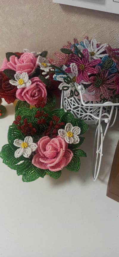 This media file features a close-up, slightly angled view of several beaded floral arrangements. The arrangement in the foreground consists of a large pink rose surrounded by green beaded leaves, with two smaller white flowers with yellow centers, and some dark red beaded accents. To the left and slightly behind this, there are more beaded flowers in shades of pink, white, and blue, many with yellow centers, cascading from what appears to be a decorative white wire basket or stand. The background is a light-colored, textured surface, possibly a wall or fabric. The lighting suggests an indoor setting with artificial light. The overall impression is of handcrafted decorative items. There are no people visible, nor any text. The location context of Asbest, Russia, does not provide any direct visual cues within the image itself.