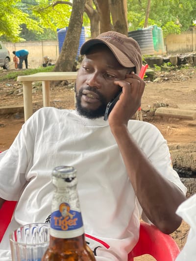 This image captures a man wearing a brown baseball cap and a white t-shirt, engaged in a phone call. He is seated outdoors, with a blurry background that suggests a casual, possibly rural setting. In the foreground, a partially visible beer bottle with a blue and yellow label, featuring the word "Tiger," is present. The man's hand is raised to his ear, holding the phone, and his expression appears thoughtful or engaged in conversation. The environment includes dirt ground, trees with green foliage, and what seem to be water tanks in the background. There are also hints of structures and possibly other people further in the distance, engaged in an activity that involves bending down near a vehicle. The lighting suggests it is daytime. The overall impression is a candid moment of communication in an open-air environment.