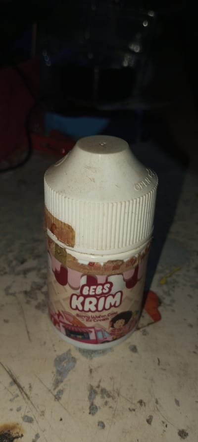 A small, cylindrical container with a white ribbed cap sits on a textured surface. The container's label reads "BEBS KRIM" and "Berry Wafer Cone - Ice Cream" with a cartoon illustration of a girl and an ice cream truck. The label is torn and peeling near the top. The white cap has "OPEN" molded into the plastic, and is dusty with a small hole on the top. The background is dark with partially visible objects.