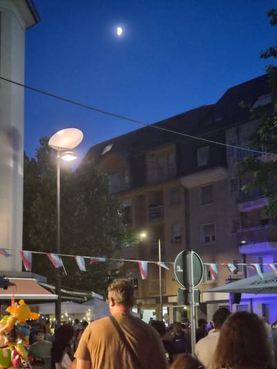 A night-time street scene is depicted, likely in Differdange, Luxembourg, with a partially visible moon in the deep blue sky. The foreground is occupied by the back of a man in a brown shirt. Behind him, a crowd of people is visible, some with their backs to the camera. To the left, a pole with plush toys is hanging. Further into the scene, a street lamp with a large disc-shaped shade illuminates the area. Buildings with multiple windows and balconies are situated in the background. Decorative flags, predominantly red, white, and blue, are strung across the street, adding to the festive atmosphere. A circular traffic sign is also visible. The overall impression is of a public gathering or event taking place at dusk or night.