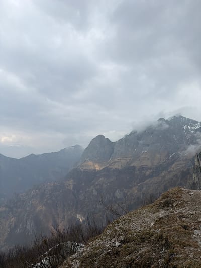 The image depicts a vast, rugged mountain landscape near Mandello del Lario, Italy, under an overcast sky. The upper two-fifths of the frame are dominated by a dense, uniform grey cloud cover. Below this, a series of rocky mountain ranges recede into the distance, exhibiting varying shades of grey and dark blue due to atmospheric perspective.
In the midground, prominent grey, craggy peaks rise, with the tallest summits to the right showing patches of snow and partially obscured by low-hanging clouds. A central mountain ridge displays areas of light yellowish-brown vegetation or dry grass, possibly illuminated by a faint light source. The lower slopes of the mountains are densely covered with dark, bare deciduous trees and dormant vegetation, indicative of late winter or early spring. Steep, rocky cliffs are visible on the far right, extending vertically. A distant, snow-capped mountain peak is faintly discernible on the far left horizon.
The immediate foreground, primarily in the lower right, consists of a steep, uneven incline composed of loose gravel, dark earth, and sparse, dry, brown herbaceous growth. Bare branches of dormant shrubs or trees are visible along the lower left edge, with small remnants of snow on the ground beneath them. No human figures, animals, or man-made objects are present in the scene.