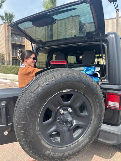 The image displays an outdoor scene in South Padre Island, United States of America, under clear daylight conditions. A black Jeep Wrangler-style SUV is parked on a light-colored paver surface. Its rear liftgate is open, angled upwards, revealing the vehicle's cargo area. A spare tire, a NEXEN ROADIAN HTX2 All-Weather Performance sized 245/75R17 on a black rim, is mounted on the vehicle's tailgate. Above the spare tire, a horizontal red LED brake light bar is present.

A female subject is partially visible on the left side, positioned behind the spare tire, facing right. She has dark hair styled in a bun with a patterned scrunchie, wears sunglasses, an orange-brown textured long-sleeved top, and a lighter undershirt. She is smiling, with her mouth open, and her right arm is extended towards the interior of the vehicle, indicating an action of loading or unloading. Inside the vehicle's cargo compartment, a folded beach chair with a blue and white pattern is visible.

In the background, multi-story residential buildings with balconies and light-colored siding are observable, along with several palm trees. A black metal fence runs between the vehicle and the background structures. The sky is light blue with minimal cloud cover.