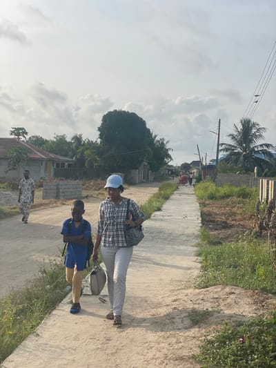 This image depicts a daytime scene in Mosafejaw, Nigeria, along a road with distinct pathways. In the foreground, a woman and a boy walk towards the viewer. The woman, positioned on a concrete pathway to the right, wears a light blue baseball cap, a white and blue plaid long-sleeved shirt, light grey trousers, and brown sandals. She carries a patterned shoulder bag on her left shoulder and a light-colored tote bag in her right hand. To her left, on an unpaved dirt road, a boy walks with his arms crossed. He wears a blue short-sleeved shirt, khaki shorts, blue sandals, and a dark backpack.
Further back on the dirt road, a man in a light-colored patterned outfit walks away from the viewer, passing a stack of concrete blocks. In the far background, several individuals, possibly students in uniform (reddish/maroon), are visible walking away on the same concrete pathway as the woman.
The environment features various low-rise buildings and dense vegetation, including palm trees, along both sides of the road. A prominent large tree stands in the mid-ground. Utility poles with wires are visible on the right side of the frame. The sky is largely overcast with white and grey clouds, indicating natural daylight conditions.