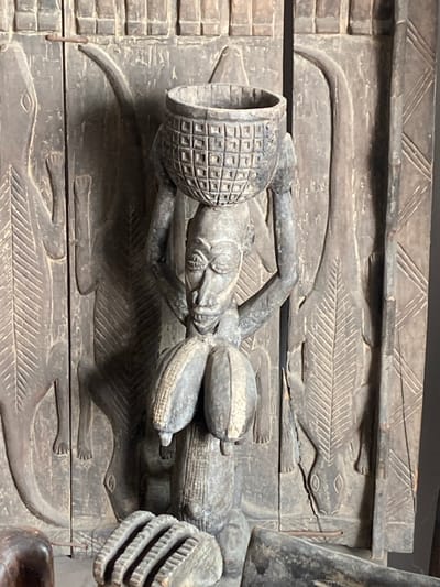 A weathered, dark brown carved wooden panel is observed in Ikate, Nigeria. The panel features a central, standing anthropomorphic figure in high relief, supporting a cylindrical basket-like object with a grid pattern on its head. The figure's face exhibits prominent, wide-set eyes, an elongated nose, and an open mouth revealing teeth. Two large, segmented, pendulous forms, resembling ears of corn, are carved on the figure's chest. This central figure is flanked by two elongated reptilian forms, possibly lizards or crocodiles, carved in lower relief, oriented vertically and facing downwards. The panel's background is textured with various carved patterns, including horizontal bands at the top and vertical stylized foliage or geometric motifs. The entire piece demonstrates characteristics of aged wood.