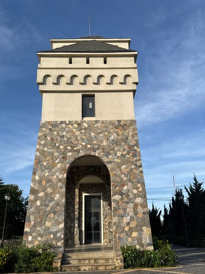 The image shows a stone-clad tower with a light-colored upper section, under a blue sky with scattered clouds. The tower has an arched entrance at its base, leading to a door. The upper section features crenellations and a small, dark window. The surrounding environment includes greenery, a lamppost, and tall evergreen trees. Power lines are visible in the background. This tower is located in Phát Chi, Vietnam.