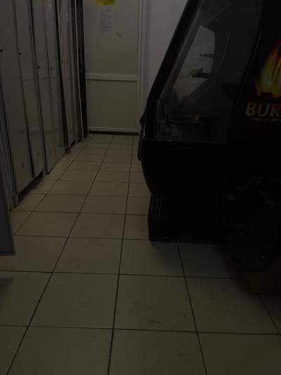 The image shows a row of metal lockers on the left and the side of a black vending machine on the right. The lockers are light gray and appear to be in a hallway or locker room. The vending machine has a logo with the word "BUR" in yellow text and flames. The floor is tiled with light-colored square tiles. The scene appears to be indoors, possibly in a public facility, with a door visible in the background behind the lockers. The lighting is dim, casting shadows. No people are visible in the image. The scene is likely located in Apsheronsk, Russia, as specified in the prompt.