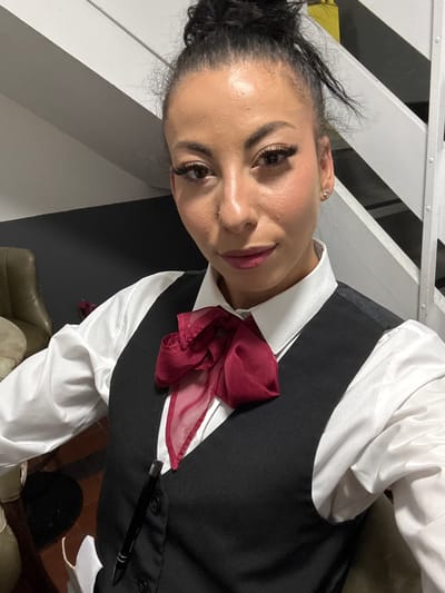 A woman with dark hair pulled back in a bun is wearing a white collared shirt, a black vest, and a red bow. She is holding a black pen clipped to her vest. The background includes white horizontal bars and dark gray areas. A tan colored chair or seating is partially visible. There is no information in the image to indicate this photo was taken in Warrington, United Kingdom.