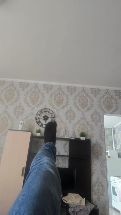 The image depicts an interior scene with a person's legs, clad in blue jeans and black socks, prominently featured in the foreground, resting on a piece of furniture. The background displays a wall with ornate wallpaper, a large circular clock with Roman numerals, a small potted plant, and a router. To the left, a section of a wooden cabinet is visible, and to the right, a portion of a kitchen area with a white microwave and stove can be seen. The scene appears to be set within a residential dwelling.