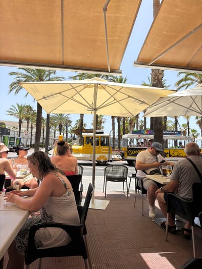 The image depicts a group of people sitting at tables under a large umbrella and awnings, likely at an outdoor cafe or restaurant. Palm trees and a bright blue sky suggest a sunny day in a warm climate, possibly a vacation destination. In the background, a yellow tourist train with advertising for "visitsalou.com" is visible, indicating the location is Salou, Spain.

Several individuals are seated at tables, with some appearing to be engaged in conversation or looking at their phones. One person is holding a phone and a glass of beer. Another person is wearing a light-colored dress with a floral pattern. The overall atmosphere appears relaxed and leisurely, consistent with a vacation setting. The time of day appears to be daytime, given the bright sunlight casting shadows. The people's attire is casual, further supporting a relaxed, outdoor environment. The image captures a moment of everyday life at a tourist spot.