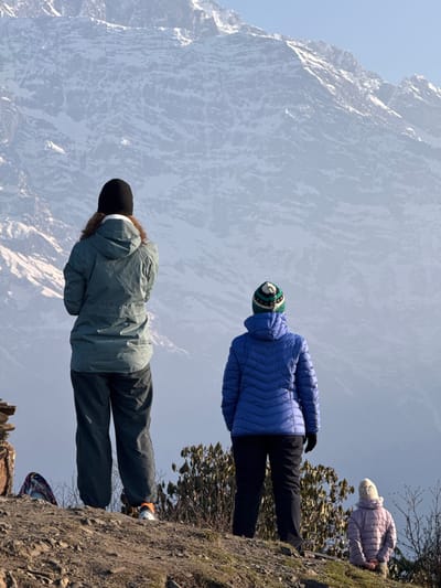 The image captures a breathtaking mountainous vista in Lwang Ghalel, Nepal, under a clear, bright sky. Three individuals, dressed in warm winter clothing, are positioned on a rocky and sparse hillside, appearing to gaze out at the majestic snow-capped peaks that dominate the background.
In the immediate foreground, a person with long brown hair, wearing a dark green or grey jacket, dark pants, and a black beanie, stands with their back mostly to the viewer, looking towards the mountains. To their right and slightly behind them, another person is visible, clad in a vibrant blue puffer jacket, dark pants, and a distinctive green, white, and blue striped beanie. Further to the right and closer to the edge of the foreground, a third individual, wearing a light purple puffer jacket and a light-colored knitted beanie, is seen sitting or kneeling on the rugged terrain, also facing the mountain range. A bag or backpack is partially visible on the ground near the front person.
The background is filled with the awe-inspiring sight of immense, rugged mountains, their peaks and upper slopes covered in snow and ice, contrasting with the clear pale blue sky. The foreground comprises dry earth and rocks, with some sparse bushes and leafless tree branches, indicative of a high-altitude environment. The bright daylight suggests it is mid-day, and the warm clothing implies cold temperatures. The scene conveys a sense of peaceful contemplation and appreciation for the natural grandeur of the Himalayas. No visible text is present.