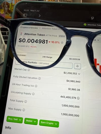 The image displays a close-up view of a smartphone screen, captured through a pair of dark-rimmed eyeglasses. The phone shows a cryptocurrency trading interface, specifically for "Attention Token" (ATTN). The price is displayed as $0.004981, with a 10.0% decrease. Other details visible include the market cap, fully diluted valuation, 24-hour trading volume, circulating supply, total supply, and max supply. There are also buttons for "Buy / Sell", "Wallet", and "Earn Crypto". The top of the phone shows the time as 10:33 and the date as Wednesday, July 1st. The background of the image is blurred but appears to be indoors, with a hint of a dog in the upper left corner and what looks like the edge of a desk or table. The image is a photograph taken by a person wearing eyeglasses, positioned to view the smartphone. There is no indication or contextual information within the image that places it in the city of Aksakovo, Bulgaria, or any specific city for that matter.