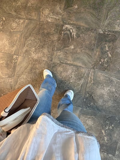 The image shows a person's legs from the knees down, standing on a tiled floor. The tiles are large and square with a gray, marbled pattern. The person is wearing blue jeans and white sneakers. They are also holding a light-colored purse with a brown textured exterior and a white zipper. The purse is resting on the person's lap. The top part of a white garment, possibly a shirt or jacket, is visible in the upper right corner of the image, with a decorative black and white trim along the edge. The lighting suggests it is daytime, with a warm glow illuminating the floor. The scene appears to be indoors, possibly in a public space like a mall, airport, or a large building lobby, given the flooring and the presence of a purse. There are no discernible emotions or specific weather conditions visible. No text is present in the image. The perspective is from above, looking down at the person's legs and the floor. The image provides a close-up view of the lower body and accessories, suggesting a casual, everyday moment.