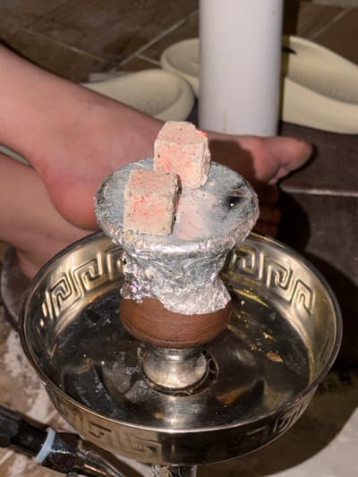 A top-down, close-up shot shows a hookah placed in the vicinity of a person's feet. The hookah features a metal base with a patterned rim, a central stem, and a bowl covered in foil. Two red-speckled charcoal cubes rest on top of the bowl. The person's bare feet are positioned near the hookah, and a white PVC pipe and light-colored sandals are visible in the background. The setting appears to be indoors, potentially on a tiled floor. There are no overt indicators to verify the location is Maafushi, Maldives.