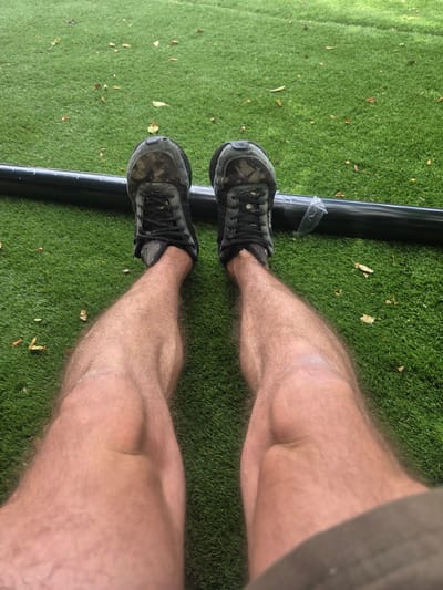 A first-person perspective shot shows a person's legs and feet, clad in black athletic shoes with camouflage patterns, resting on artificial turf. A black pipe runs horizontally across the image, with a clear plastic wrapper partially covering it. The artificial turf is scattered with fallen leaves, suggesting an outdoor location. The scene is set in Elmbridge, United Kingdom.