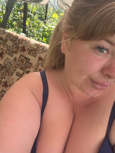 The media file contains a close-up, low-angle, portrait-style photograph of a young woman lying down outdoors. She has fair skin with freckles and light brown hair. Her eyes are green and her lips are slightly parted. She is wearing a dark blue bikini top. The background shows lush green foliage, suggesting it is daytime and likely warm weather. The woman is positioned on a patterned fabric surface, possibly a cushion or blanket, which is resting on an outdoor seating structure. The image is oriented sideways. There is no visible text. The overall mood is relaxed and casual. The location is identified as Kimry, Russia.