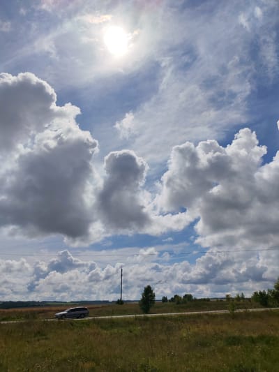 The image depicts a landscape scene under a cloudy sky with the sun partially obscured by clouds. In the foreground, there is a grassy field with dry vegetation. A paved road runs horizontally across the middle ground, and on this road, a silver station wagon is traveling from left to right. Beyond the road, the landscape consists of fields with some trees and vegetation. In the background, there is a distant horizon with more trees. A tall, dark pole, possibly a utility pole or a radio tower, stands to the right of the road. The sky is filled with large, puffy white and gray cumulus clouds, with patches of blue sky visible between them. The sun appears as a bright, white circle peeking through the clouds in the upper left quadrant of the image, casting a soft light on the scene. This scene is located in the vicinity of Belka, Russia.