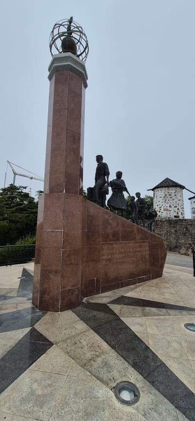 The image shows a monument with sculptures of people at its base, located in Póvoa de Varzim, Portugal. The monument is tall and rectangular, made of dark red granite, and topped with a globe-like structure. At the base of the monument, bronze sculptures depict a family, including a man, a woman holding a baby, and several children. The sculptures are positioned on steps leading up to the monument. An inscription is visible on the side of the monument. In the background, two old stone windmills with conical roofs are visible, along with lush green trees and a construction crane in the distance. The ground in front of the monument is paved with geometric patterns of light and dark stone tiles. The sky is overcast.