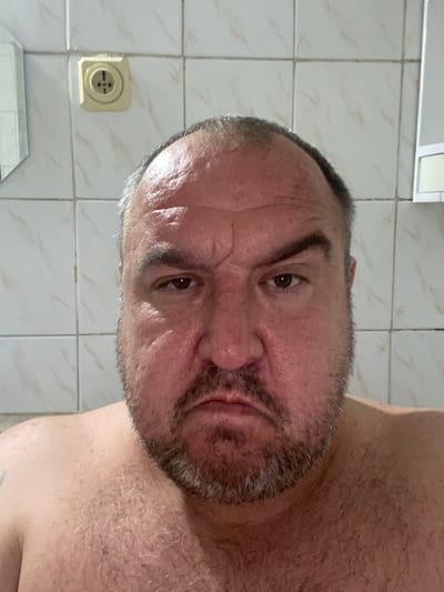 The image is a close-up, frontal, waist-up shot of a man with a beard and short hair. He has fair skin with rosy cheeks and visible wrinkles on his forehead. His expression is somber, with a furrowed brow and downturned lips. He is shirtless, revealing hairy chest and shoulders. In the background, there are light-colored tiled walls and a cream-colored electrical outlet. The scene appears to be indoors, possibly a bathroom, given the tiling and outlet. The location in Liutizh, Ukraine is not visually evident in the image.