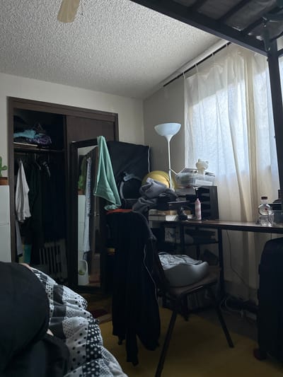 This image depicts an indoor scene, likely a bedroom, with a desk and a closet. The room is somewhat cluttered, with clothes hanging in the closet and various items on the desk. A floor lamp is positioned near the desk, and sheer curtains cover a window. A bed is partially visible in the foreground, covered with a patterned comforter. The overall impression is of a lived-in space. The image does not contain information that would allow for determining its location within the city of Vancouver, United States of America.