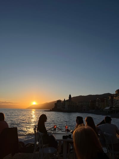 The image captures a sunset over the sea at Camogli, Italy, featuring silhouettes of people seated at outdoor tables by the water's edge. The sun, positioned low on the horizon, casts a golden glow on the water and the coastline. Buildings and a mountain range line the coastline, creating a backdrop to the scene. Cocktails are visible on the tables, and the silhouettes of people suggest a relaxed atmosphere.