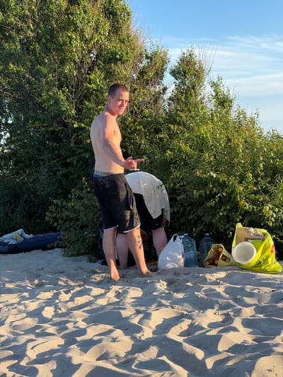 A young man stands on a sandy beach in Povarovka, Russia, facing away from the camera and looking towards a dense green bush. He is shirtless and wearing black shorts. Another person, also shirtless and wearing a light-colored garment over their lower body, is crouched down near the bush, seemingly engaged in an activity. In the foreground, the sand is textured with shadows from the setting sun, and various items are scattered on the beach, including plastic bags, large water bottles, and a yellow bag with a white bowl inside. The sky above is a clear blue with a few wispy clouds.