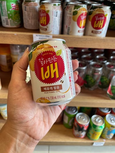This image depicts a human hand holding a beverage can within a retail store environment in the city of Anápolis, Brazil.

**Scene Description:**
The foreground is dominated by a medium-toned human hand, exhibiting light pink polished fingernails, actively holding a white beverage can. The can's label is primarily red with large yellow Korean characters reading "갈아만든 배" (Grinded Pear). Below this, "PEAR PUREE 12%" is printed in English. Additional text includes "SINCE 1996" and "238 mL (116 Kcal)". The design also incorporates illustrations of pears and leaves.

The background consists of light brown wooden shelves, typical of a retail display. The upper shelf holds multiple identical "갈아만든 배" cans, neatly arranged. A white price or product label on this shelf is partially visible, displaying "REFRESCO MISTO" (Mixed Refreshment) and "PERA ASIÁTICA" (Asian Pear), affirming the product's type and origin. Other green-labeled cans are also present on this shelf. The lower shelf, positioned below the primary subject, displays an assortment of other beverage cans, including one featuring an anime character (Goku from Dragon Ball) and others with colorful green and yellow designs. The entire scene is brightly illuminated, consistent with an indoor commercial setting.