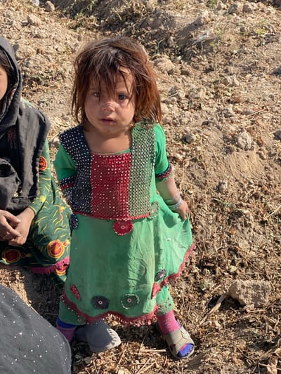 An image captured in Mandisar, Afghanistan, depicts a young girl standing in the center-right, looking directly at the viewer. She possesses reddish-brown, slightly disheveled hair that partially obscures her forehead. Her face is smudged beneath the nose. She wears a long-sleeved green dress featuring a square neckline; the bodice is vertically striped with sections of black polka dots, red embellishments, and green embellishments. The skirt portion of the dress is plain green with red lace trim and circular fabric appliques in red, black, and green. Visible beneath the skirt are green trousers with red trim, and multi-colored socks or leggings (pink and blue). She wears a grey slip-on shoe on her left foot and a tan/green sandal with a floral emblem on her right foot. Multiple silver-colored bangles adorn her right wrist, and her right hand gently holds the fabric of her skirt. To the left and slightly behind her, a portion of another individual is visible, wearing a dark green patterned garment with red and orange motifs, and a black head covering. Their right hand is visible, resting with fingers partially interlaced. The ground is dry, light brown, loose soil mixed with numerous small rocks, pebbles, and sparse dried vegetation, under bright daylight conditions.