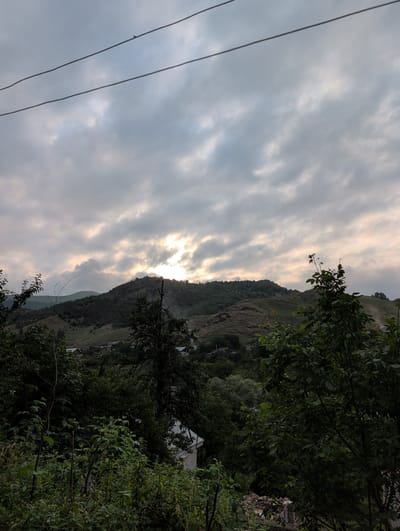 The image shows a view of a hillside village in Toğanalı, Azerbaijan, under a cloudy sky. In the foreground, dense green foliage and trees obscure the lower part of the scene, with glimpses of rooftops and structures belonging to the village. The middle ground features rolling hills covered in vegetation, with a cluster of houses and buildings visible on the slopes. The background is dominated by a larger, more rugged mountain range, partially illuminated by sunlight breaking through the clouds. Overhead, power lines stretch across the frame. The overall impression is one of a rural, mountainous landscape at dusk or dawn.