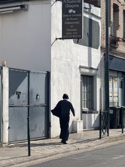 The scene depicts a daytime street in Asnières-sur-Seine, France, identifiable by the commercial sign for "A Domicile Glaçons" located at what appears to be 30, Rue des Bourguignons. A lone male pedestrian, viewed from behind, is walking on a cobblestone sidewalk from the right towards the left side of the frame. He wears a brown cap, a dark gray long-sleeved top over a lighter shirt collar, dark pants, and dark shoes, carrying a dark bag or garment draped over his left shoulder.
To the left of the pedestrian, a large, dark gray double metal gate with two visible latch mechanisms is set into a white stucco building. To the pedestrian's right, a vertical dark gray commercial sign is mounted on the white stucco building, displaying "A Domicile Glaçons" with an ice cube graphic, advertising "Vente et Livraisons de Glaçons Professionnels et Particuliers," along with business hours (24h/24 7J/7) and a phone number (01.47.98.47.93). Below the sign and to the right of the pedestrian, a window with visible vertical bars is present in the white wall. Further right, a section of a brick building is partially visible.
The cobblestone sidewalk transitions to an asphalt road in the foreground. A dark green municipal bollard is positioned in the lower-left foreground, and a dark green metal fence runs along the right side of the sidewalk. Two dark green rectangular trash bins are located on the sidewalk further right. The environment is brightly illuminated by sunlight, creating sharp shadows from the structures and the pedestrian.