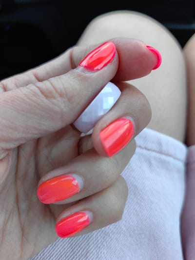 A close-up, indoor shot focuses on a person's hand with brightly colored neon orange nails. The person is holding a faceted white ring. The background is out of focus, showing what appears to be light-colored fabric, possibly clothing. The lighting is soft, and the image is sharply focused on the hand and nails, suggesting a personal or product-focused photograph. The location is not specified in the image or context.