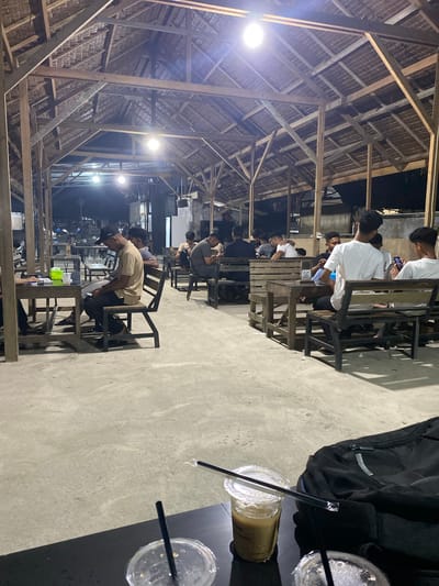 The image captures an indoor or semi-outdoor gathering place at night in Seuneubok Teungoh, Indonesia. The scene is illuminated by several bright lights, suggesting evening hours. The structure features a distinctive wooden beam framework and a thatched roof, giving it a rustic or traditional aesthetic.
Numerous individuals, predominantly men, are seated at wooden tables and chairs arranged in rows. Many appear to be engaged with their mobile phones. One person in the foreground, wearing a light-colored shirt and a dark baseball cap, is leaning forward on the table.
In the immediate foreground, on a dark table, there is a black padded backpack. Beside it, three plastic cups are visible; one contains a light brown beverage, possibly iced coffee, with a straw, while the other two appear mostly empty with straws. The table surface is a concrete or light-colored floor.
A notable detail is a white card with the number "16" printed on it, visible on one of the wooden tables in the midground. The overall atmosphere appears casual and social. The image is oriented vertically, rotated 90 degrees clockwise from its natural horizontal perspective.