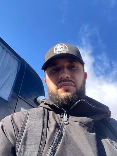 A man in a black baseball cap and jacket is taking a selfie outdoors, with a bright blue sky and some clouds in the background. A black bus is visible on the left side of the image. The man has a beard and is looking directly at the camera. The image appears to be taken during the day.