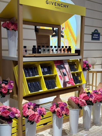 The image depicts a retail display featuring Givenchy perfumes. The display is brightly lit with a yellow overhead sign bearing the "GIVENCHY PARIS" logo. Various bottles of perfume are arranged on shelves, with some displayed prominently on a wooden shelf in the upper section and others in yellow bins below. The lower shelves are filled with boxed perfume sets. Decorative elements, including bouquets of pink and purple artificial flowers in white vases, are strategically placed around the display, both on higher shelves and at the bottom. In the background, a sign on a wall reads "8° Arr. AVENUE GEORGE V," suggesting a Paris street address. The overall environment appears to be an indoor retail space, likely a department store or a dedicated brand boutique, with a light-colored tiled floor and white paneled walls. There are no people visible in the image. The scene is clean and well-organized, highlighting the products and the brand's aesthetic. The location is not Arnavutköy, Turkey; the sign clearly indicates an address in Paris, France.