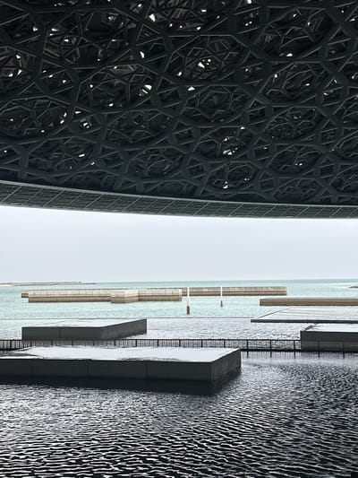 The image shows a section of the Louvre Abu Dhabi in Abu Dhabi, United Arab Emirates. On the left is a portion of the museum's dome, which features an intricate, geometric pattern of overlapping metal structures. To the right of the dome is a light blue expanse of water that appears to be the sea, with several elevated concrete structures and walkways extending over it. The water surface shows distinctive wave patterns, creating a textured effect. The sky is a uniform pale gray, suggesting overcast conditions. There is no indication of people or interaction within the frame.