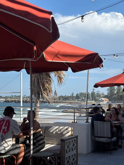 A partially-obstructed view from an outdoor patio reveals red umbrellas and a glimpse of the ocean. Patrons sit at tables, enjoying the scenery. A palm tree provides a tropical touch. The sky is blue with fluffy white clouds. A string of lights hangs overhead. The scene suggests a relaxed atmosphere at a seaside restaurant.