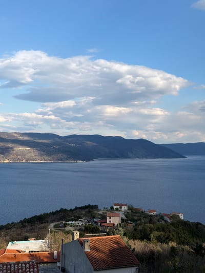 The scene depicts a view of Zagorje, Croatia, with a bright blue sky and fluffy white clouds on the left side of the frame. Moving rightward, a rugged mountain range with varying shades of green and brown flanks a body of water, likely the Adriatic Sea. The right side of the image showcases a coastal landscape with houses clustered along the hillside, intermingled with trees and vegetation. Some rooftops are visible in the foreground, suggesting the vantage point is from a higher elevation.