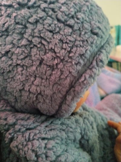 A close-up shows a person wearing a gray, hooded, sherpa-like jacket. The hood is up, covering the head, and the texture of the material is prominent. The person is also holding a finger. The background is blurry.